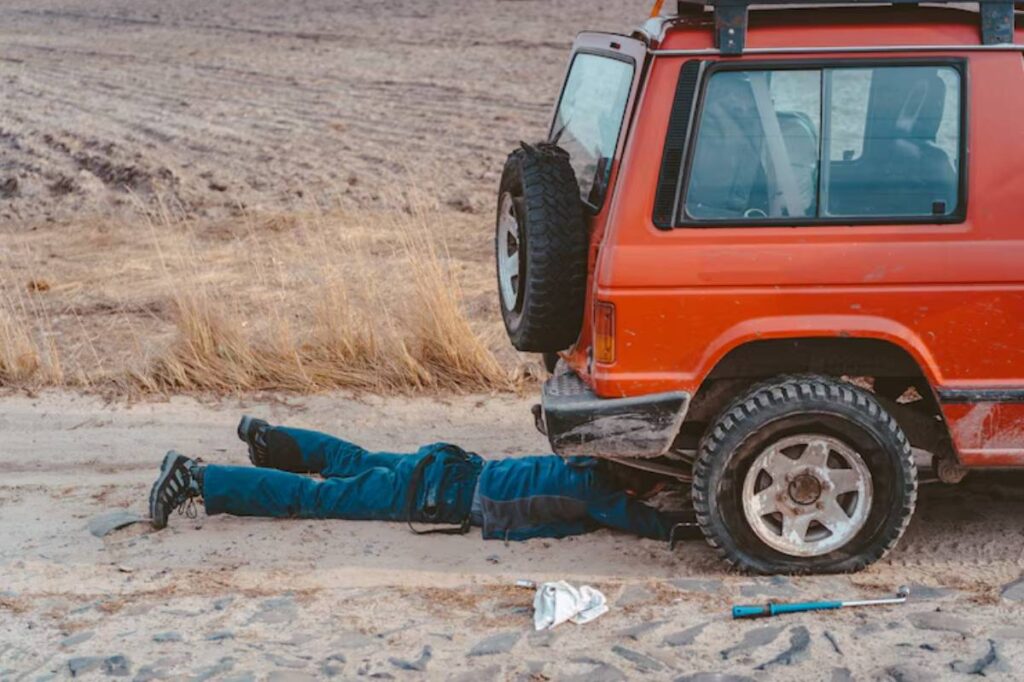 A person in blue coveralls lies under an orange SUV, working on it, with tools and a partially sandy ground around them.