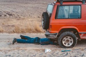 A person in blue coveralls lies under an orange SUV, working on it, with tools and a partially sandy ground around them.