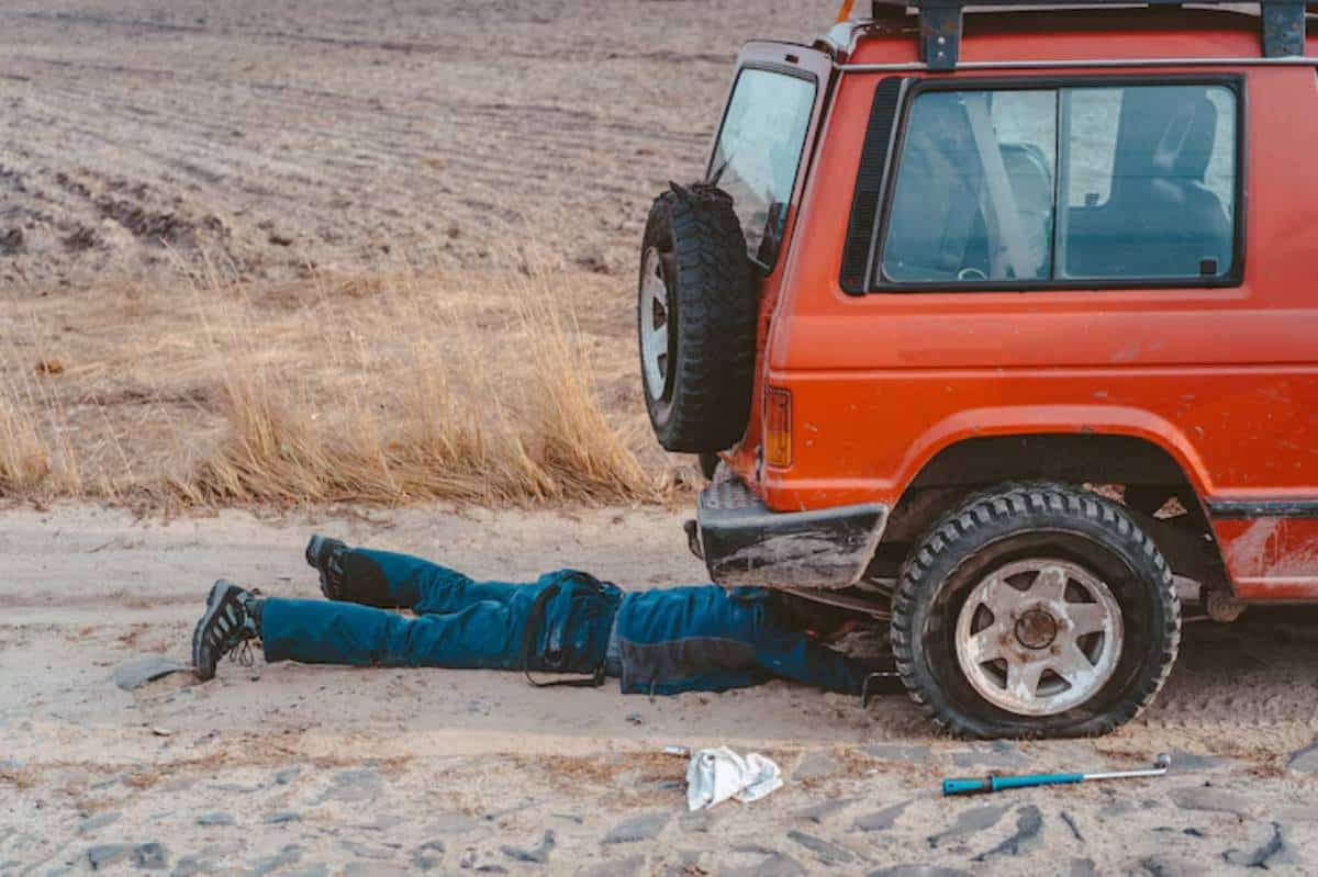 A person in blue coveralls lies under an orange SUV, working on it, with tools and a partially sandy ground around them.