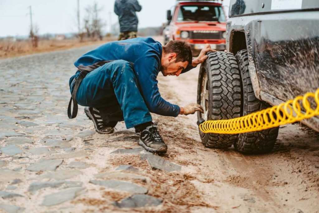 A person crouches beside a vehicle on a gravel road, using tools to inspect or fix a tire, with a second vehicle in the background.