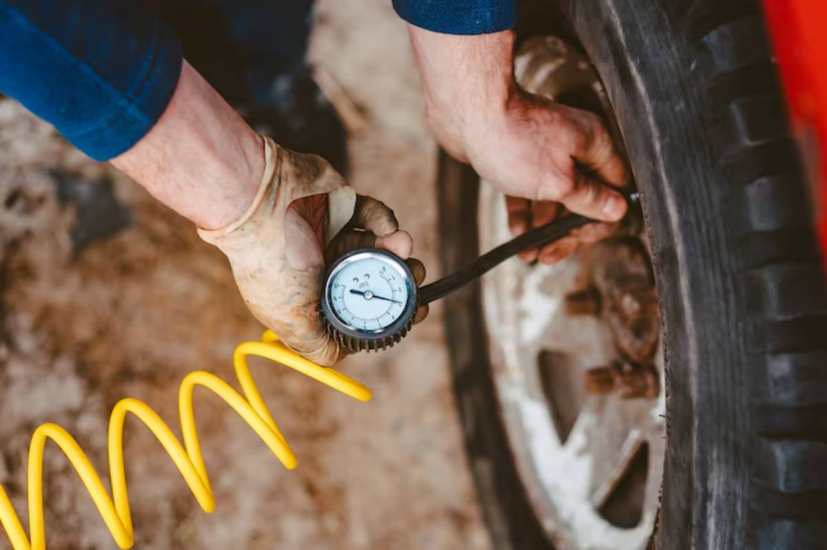A mechanic checks tire pressure using a gauge, with a yellow air hose coiled nearby, against a muddy background.