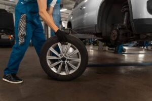 A mechanic in blue overalls lifts a shiny alloy wheel in a garage, preparing to install it on a vehicle on a lift.