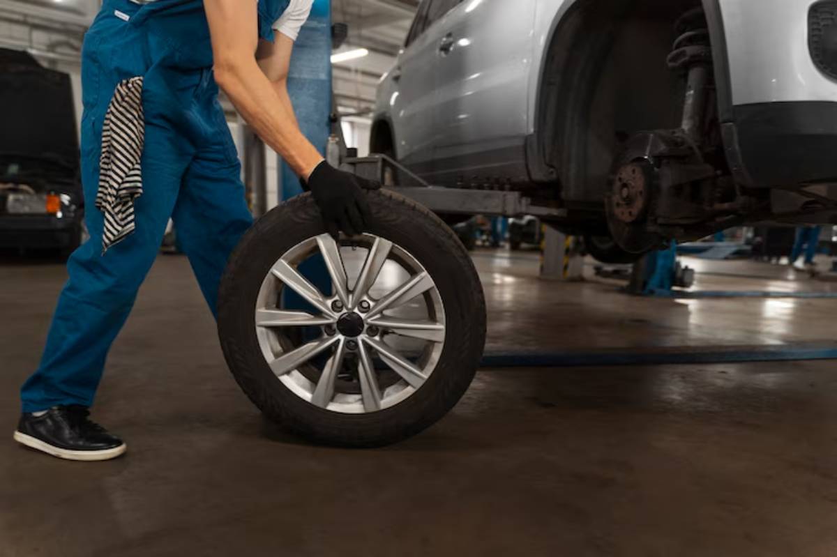 A mechanic in blue overalls lifts a shiny alloy wheel in a garage, preparing to install it on a vehicle on a lift.