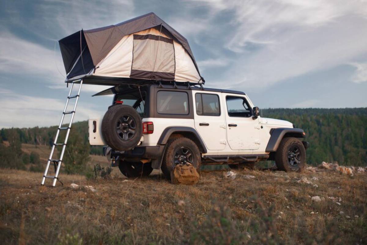 A white off-road vehicle with a rooftop tent and a ladder parked on a hillside, surrounded by greenery and under a blue sky.