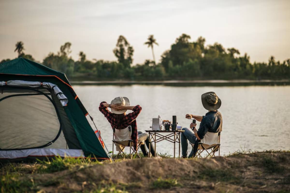 Two campers relax by a river, enjoying the view while seated by a portable table, with a tent nearby and lush greenery surrounding them.