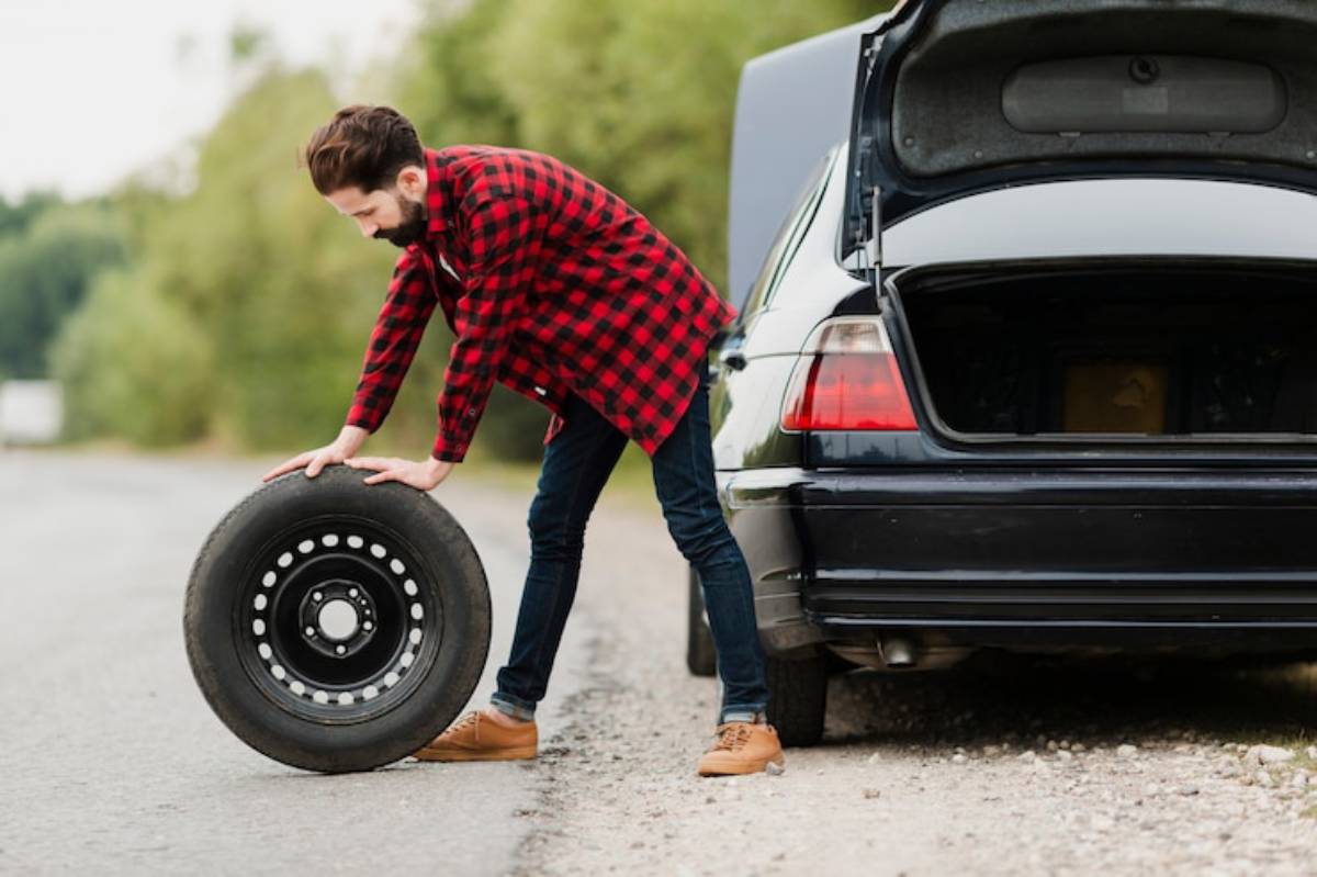 A person in a red checkered shirt is lifting a car tire next to a parked black vehicle with its trunk open on a gravel road.