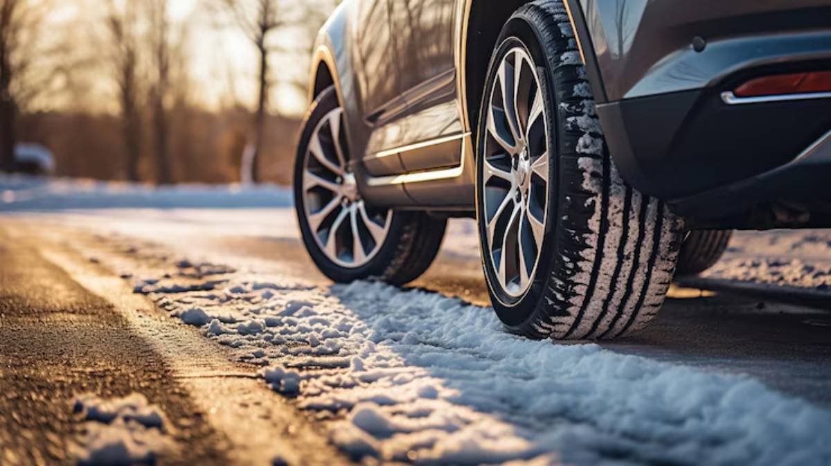 Close-up of a car tire on a snowy road, with sunlight illuminating the scene and trees in the background.