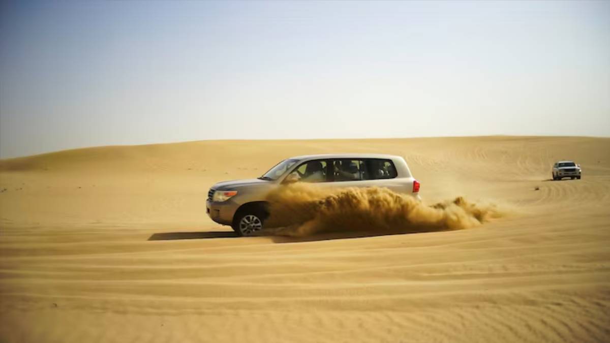 A silver SUV speeds through a sandy desert, kicking up dust as another vehicle follows in the background under a clear blue sky.