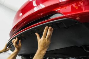 A mechanic's hands install a protective undercarriage panel on a red car in a garage, showcasing automotive maintenance.