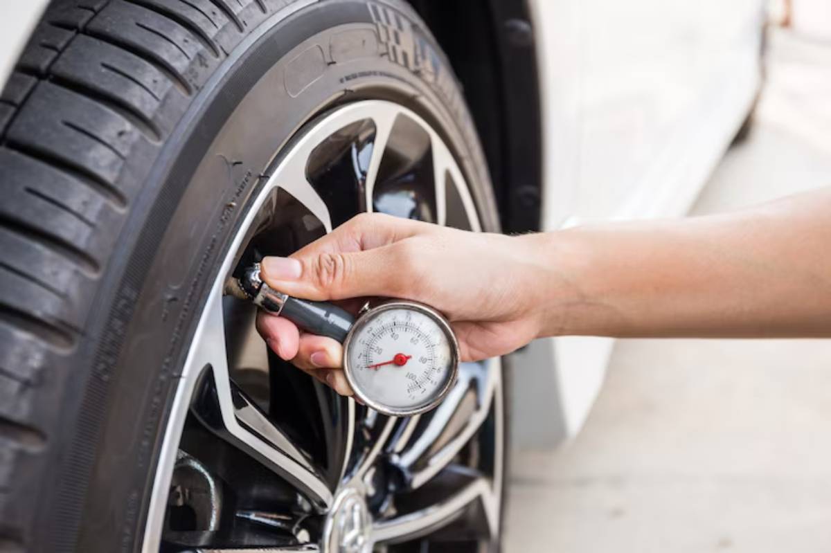 A hand holds a tire pressure gauge against a car tire, checking air pressure on a sunny day. Black alloy wheels are visible.