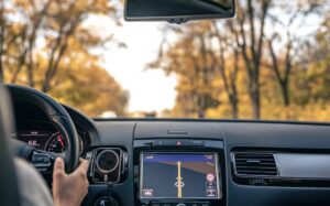 A driver's hand grips the steering wheel, while a GPS screen displays directions against a backdrop of autumn trees.