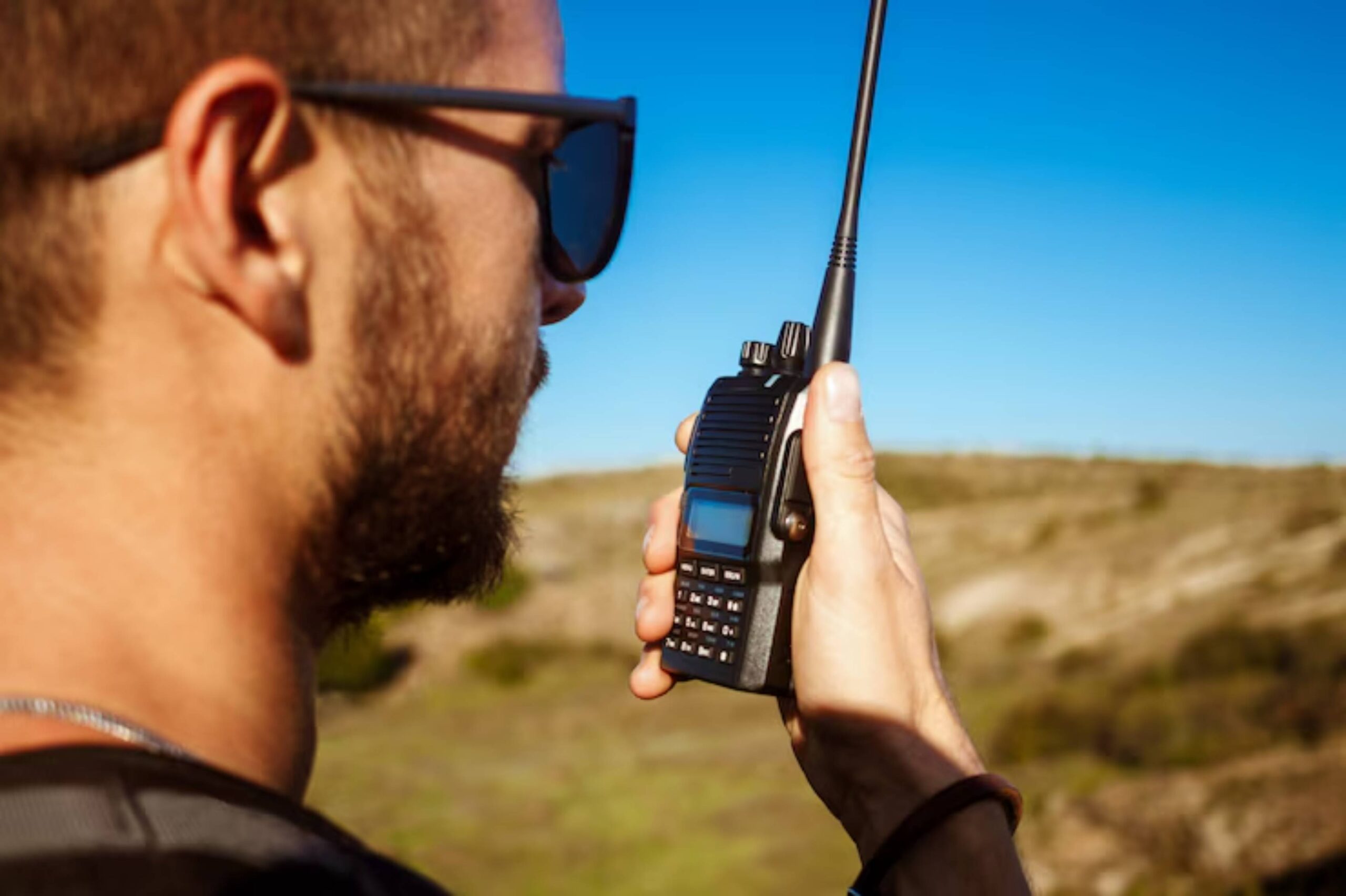 A person holds a two-way radio while standing outdoors, surrounded by a hilly landscape under a clear blue sky.